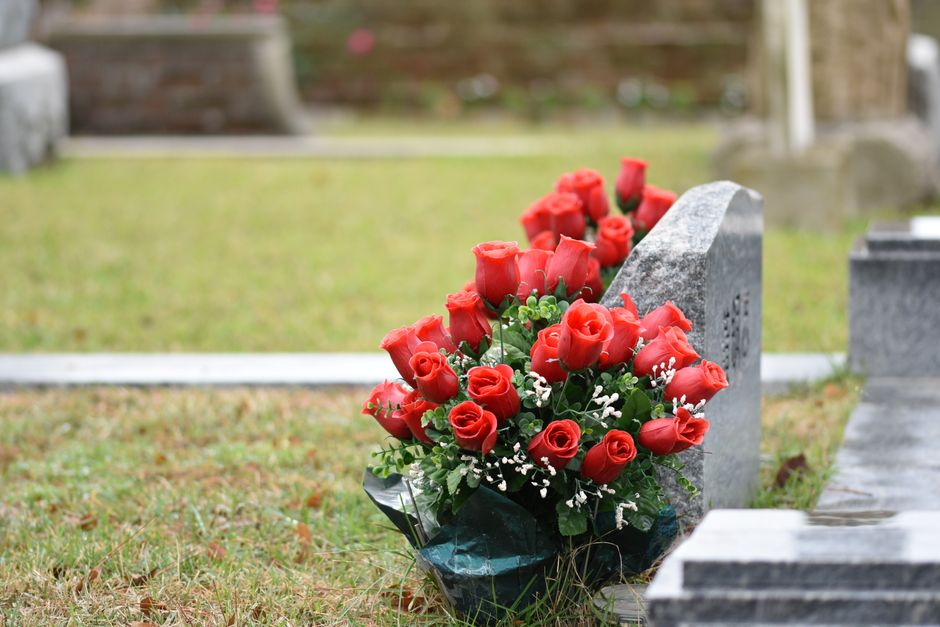 A bouquet of vibrant red roses, arranged with small white baby's breath flowers and lush green foliage, is placed against a grey granite memorial stone in a cemetery. The flowers are wrapped in dark g