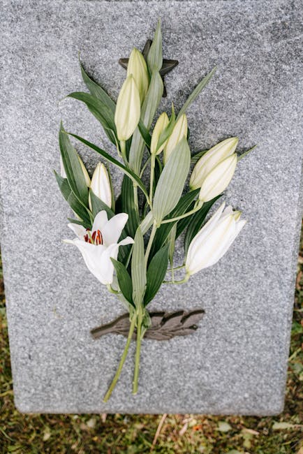 A memorial flower arrangement attached to a grey granite memorial stone, featuring white lilies with large blooms and elongated green leaves, complemented by several white tulips with closed buds and 