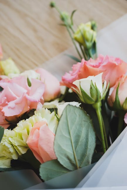 A close-up view of a floral arrangement featuring light pink and cream roses with soft, ruffled petals and fresh green leaves, arranged in a bouquet with some flowers still in bud form. The bouquet is