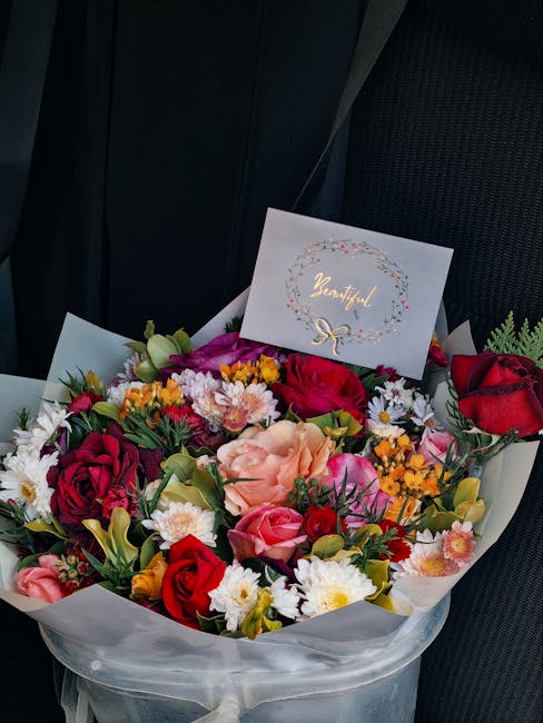 A vibrant floral arrangement featuring a mix of red, pink, and peach roses, complemented by white daisies, purple chrysanthemums, and sprigs of yellow and orange flowers, all arranged in a rounded bou